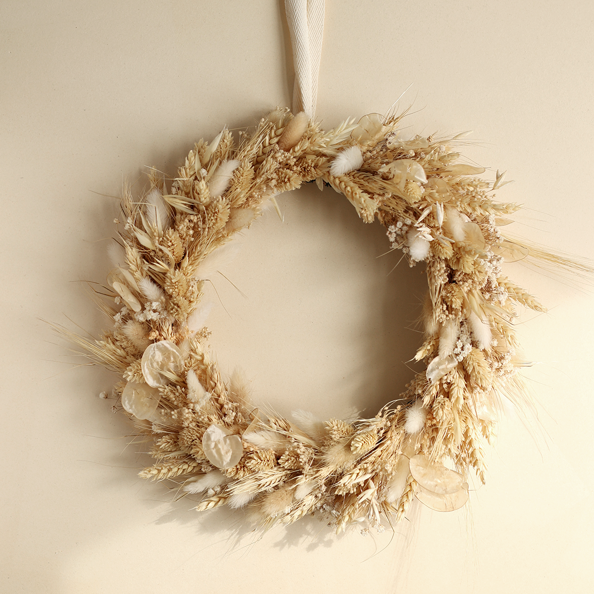 Ein handgebundener Flowerhoop aus hellen Trockenblumen, Gräsern und Getreideähren, der an einem cremefarbenen Band vor einer neutralen Wand im Wiener Studio hängt.