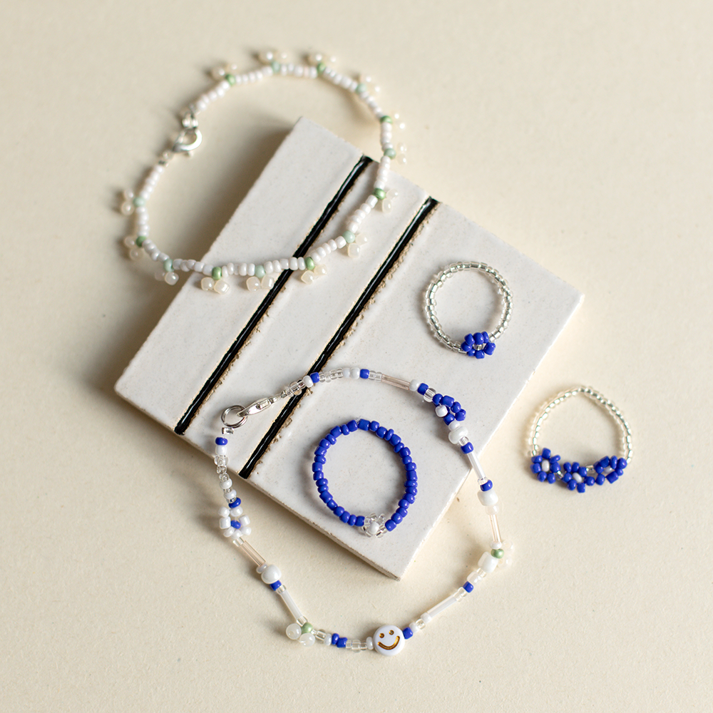 Selbstgemachter Perlenschmuck, bestehend aus Armbändern und Ringen in Blau- und Weißtönen mit Blumenmustern und Smiley-Details, präsentiert auf einer Keramikfliese.
