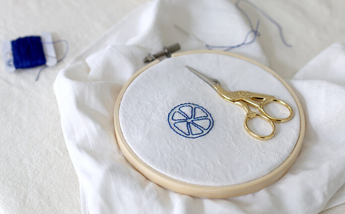 Stickrahmen mit einer blau gestickten Zitronenscheibe auf weißem Stoff, daneben eine goldene Storchenschere und eine blaue Stabkerze – Fokus auf achtsames Handarbeiten und Selfcare. | we love handmade
