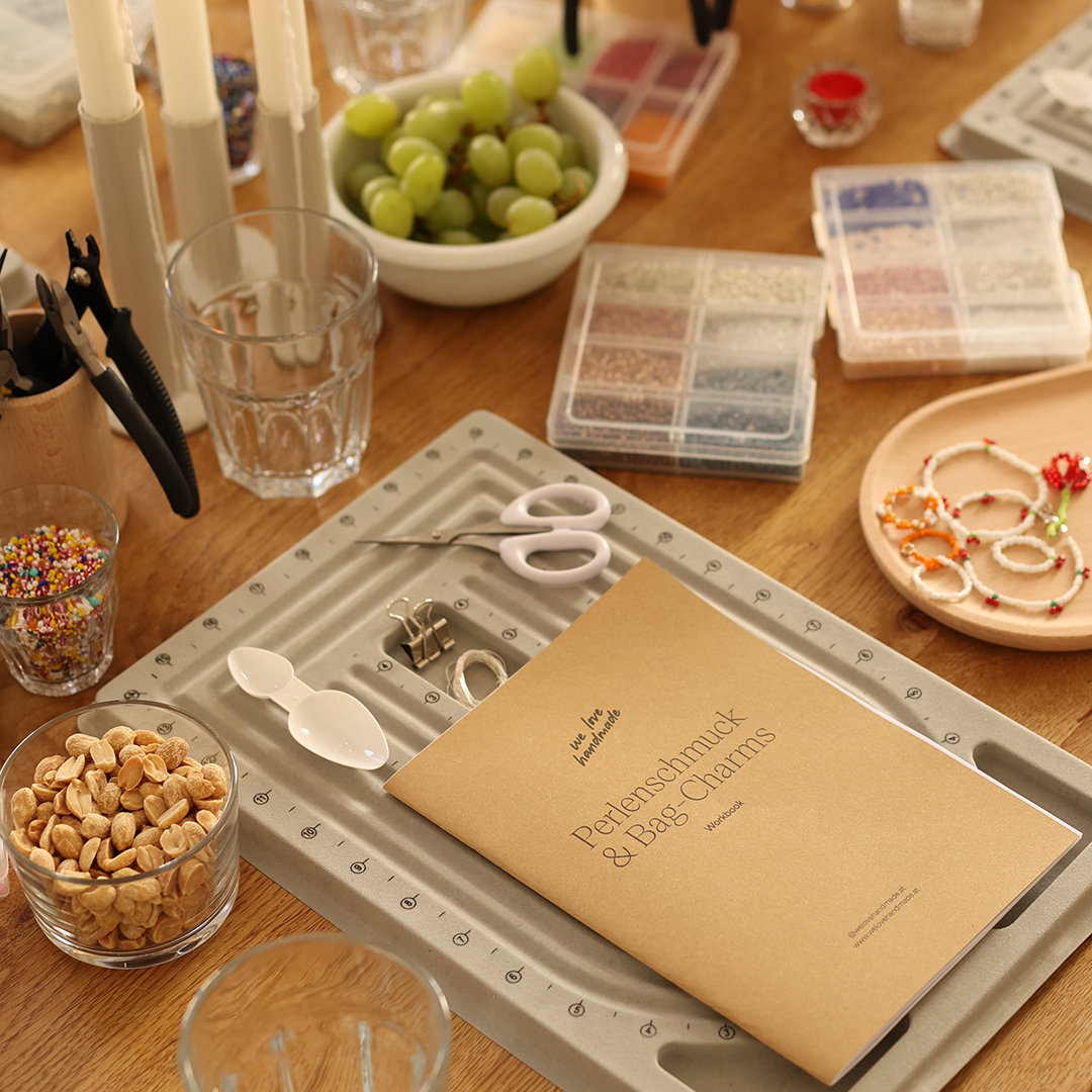 Ästhetisch gedeckter Workshop-Tisch mit Perlenbrettern, Zangen, dem we love handmade Perlenschmuck-Workbook, Weintrauben und Knabbergebäck.