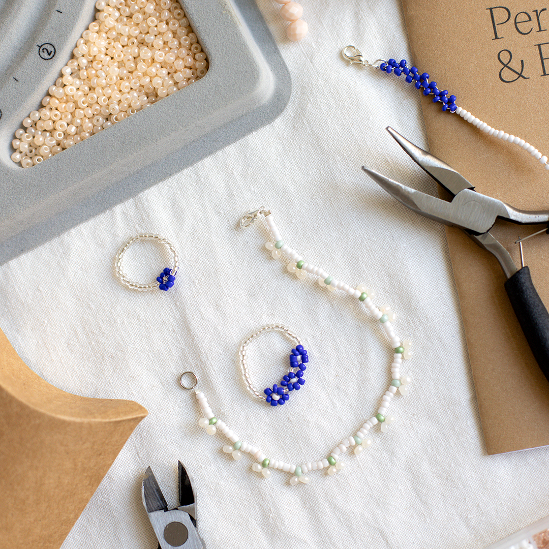 Eine saubere Draufsicht auf Perlenboxen, Schmuckzangen, eine Perlenschale und fertige Perlenarmbänder auf einem hellen Leinentuch im we love handmade Studio.