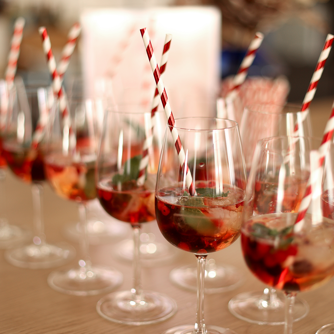 Eine Reihe eleganter Weingläser gefüllt mit Aperitivo, Eiswürfeln, frischen Beeren, Minze und rot-weiß gestreiften Papierstrohhalmen auf einem Holztisch bei einem Event. | we love handmade