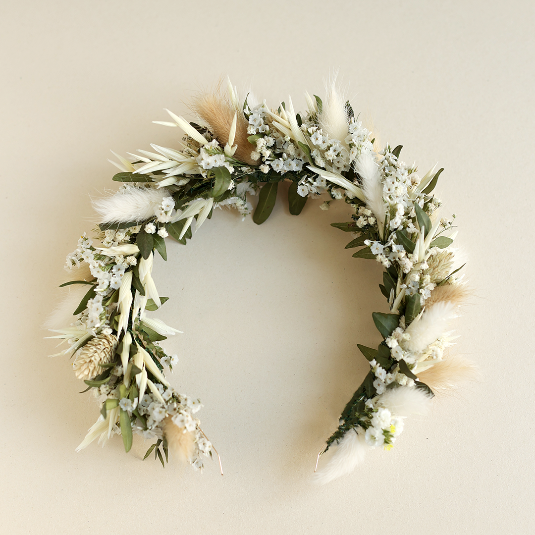 Ein handgemachter Haarreif-Blumenkranz von we love handmade, gebunden aus frischen grünen Pistazienzweigen, weißen Trockenblumen, Schleierkraut und flauschigem Samtgras auf beigem Hintergrund.