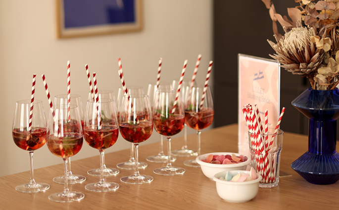 Eine Reihe gefüllter Aperitivo-Gläser mit Beeren und rot-weiß gestreiften Strohhalmen auf einem Holztisch im we love handmade Studio in Wien Neubau. | we love handmade