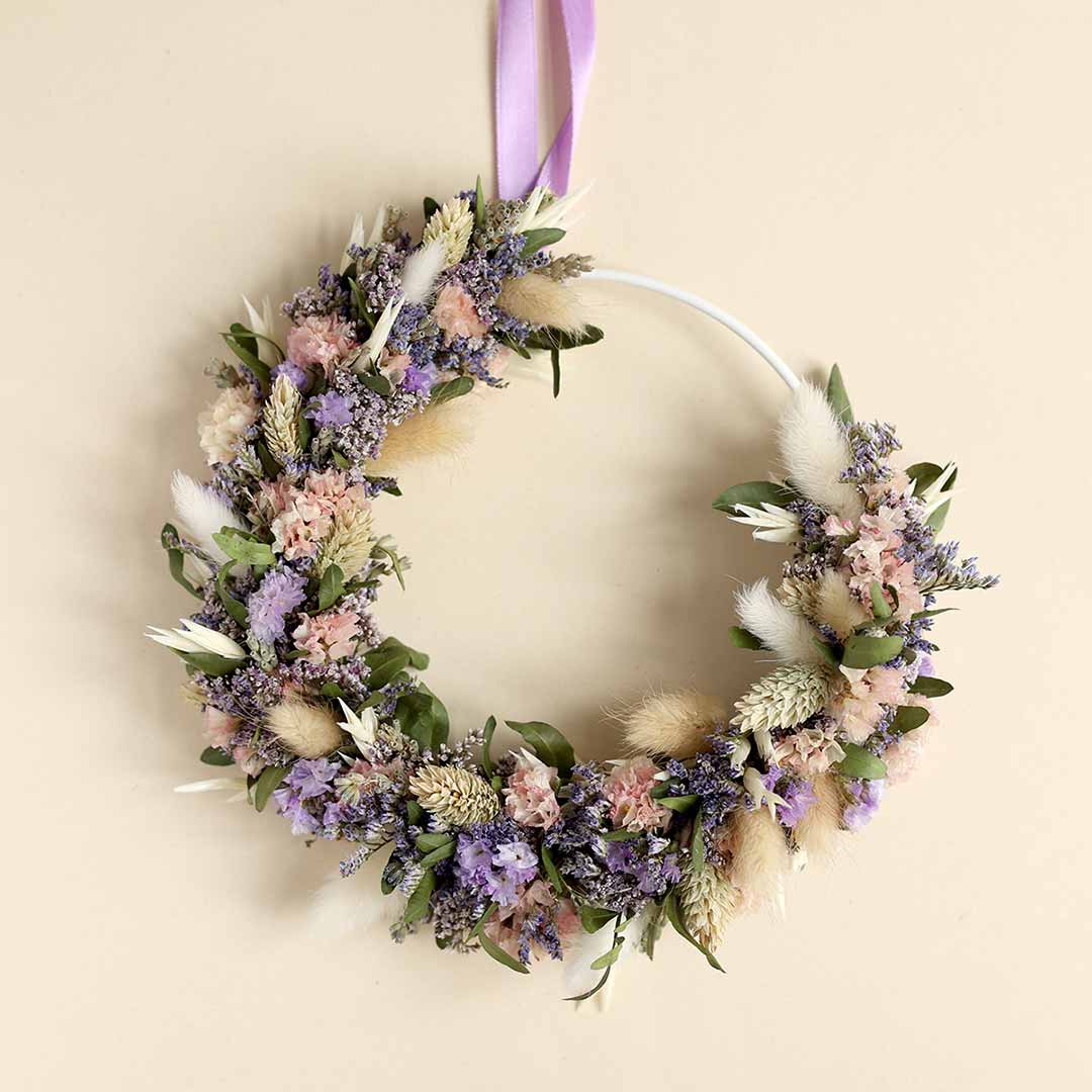 Ein moderner Flowerhoop auf einem weißen Metallring, gebunden mit Trockenblumen in Rosa, Lila und Naturtönen, hängend an einem lila Satinband vor neutralem Hintergrund.