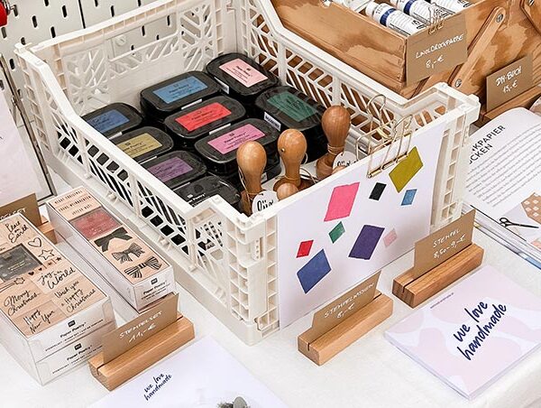 Nahaufnahme eines weißen Klappkastens voll mit Stempelkissen und Stempeln auf dem we love handmade Stand. Dahinter Holzboxen mit Farben und Flyer für DIY-Workshops.
