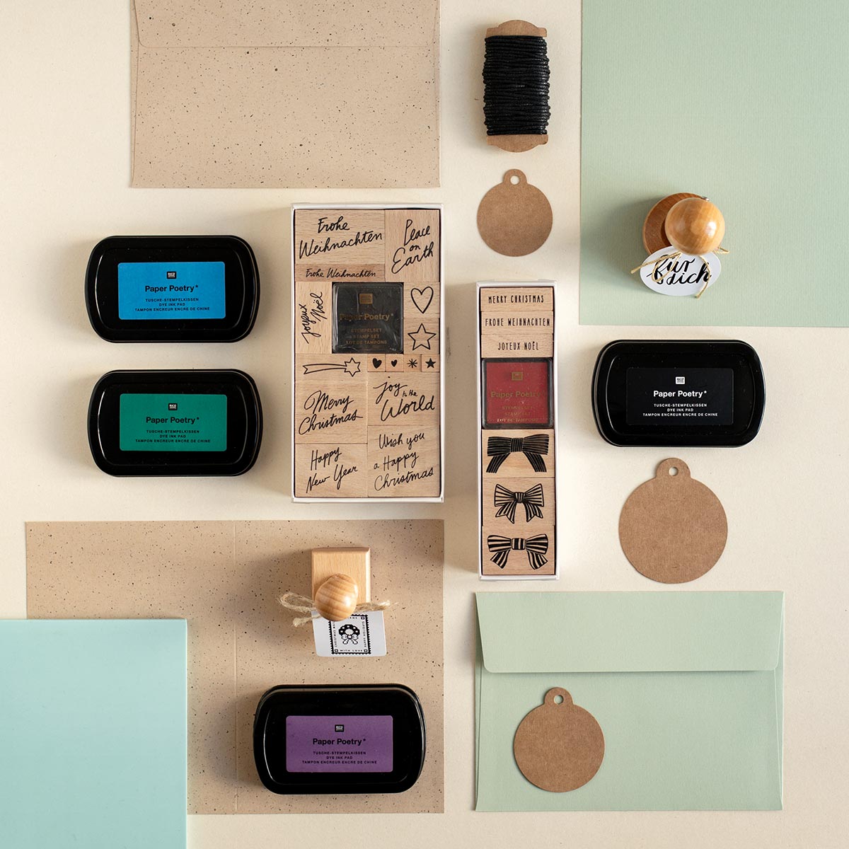 Flatlay Aufnahme von Paper Poetry Bastelbedarf auf beigefarbenem und mintgrünem Papier. Zu sehen sind Stempelkissen in Blau, Grün, Violett und Schwarz, zwei Holzstempel-Sets mit Weihnachtsmotiven und Schriftzügen, Geschenkanhänger aus Kraftpapier, Kordel auf Spule und Briefumschläge.